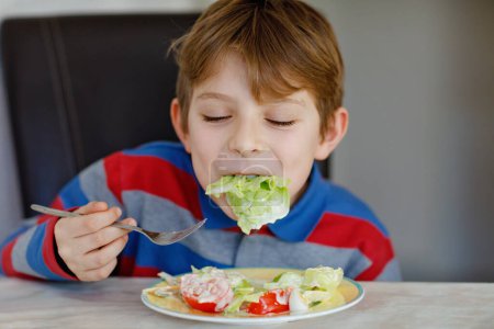 Foto de Niño feliz comiendo ensalada fresca con tomate, pepino y diferentes verduras como comida o merienda. Niño sano disfrutando de comida sabrosa y fresca en casa o en la cantina de la escuela - Imagen libre de derechos