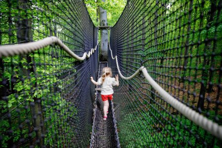 Photo for Cute little preschool girl walking on high tree-canopy trail with wooden walkway and ropeways on Hoherodskopf in Germany. Happy active child exploring treetop path. activity for families outdoors. - Royalty Free Image