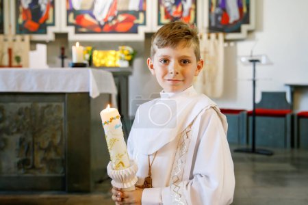 Petit garçon recevant sa première sainte communion. Joyeux enfant tenant une bougie du baptême. Tradition en curch catholique. Enfant en robe traditionnelle blanche dans une église près de l'autel

