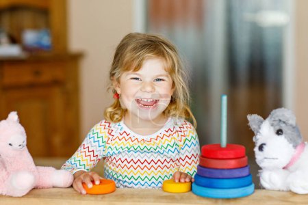 Cute little toddler girl playing alone with colorful wooden rainbow pyramid and toys at home or nursery. Happy healthy child having fun in kindergarten or preschool daycare.