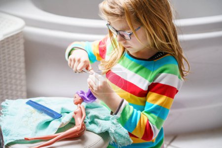 Photo for Cute little girl playing with doll at home. Child with eyeglasses making haircut to her toy doll. Indoor creative activities for children. - Royalty Free Image