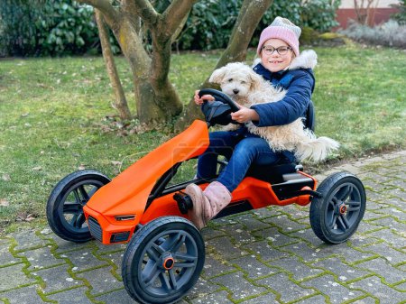 Téléchargez les photos : Souriant heureuse écolière avec chien maltais s'amuser sur la voiture à pédale, à l'extérieur. Enfant et famille animal de compagnie ensemble - en image libre de droit