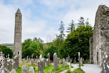 Foto de Torre redonda de piedra y algunas ruinas de un asentamiento monástico construido originalmente en el siglo VI en el valle de Glendalough, Condado de Wicklow, Irlanda en un día soleado
. - Imagen libre de derechos