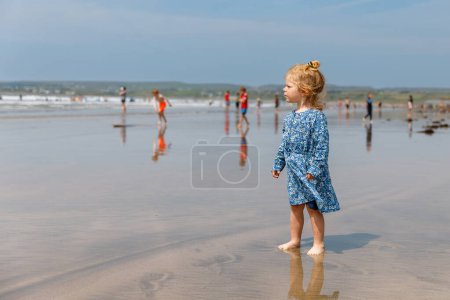 Foto de Pequeña niña linda en la playa de surfistas Ballybunion, divirtiéndose jugando en la costa oeste de Irlanda. Niño feliz disfrutando del verano irlandés y el día soleado con la familia - Imagen libre de derechos