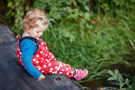 Photo for Cute adorable toddler girl sitting on wooden bridge and throwing small stones into a creek. Funny baby having fun with outdoor games in nature. Active outdoors leisure and activity with little kids - Royalty Free Image