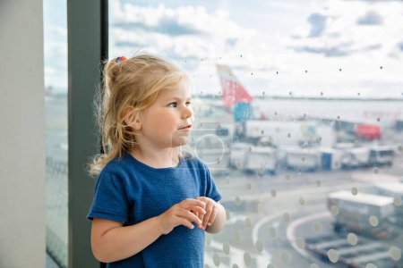 Foto de Linda niñita en el aeropuerto, viajando. Feliz niño sano esperando cerca de la ventana y viendo aviones. Familia de vacaciones de verano en avión - Imagen libre de derechos