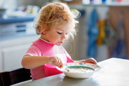 Adorable baby girl eating from spoon vegetable noodle soup. Healthy food, child, feeding and development concept. Cute toddler child with spoon sitting in highchair and learning to eat by itself.