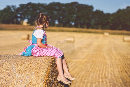 Cute little kid girl in traditional Bavarian costume in wheat field. Happy child with hay bale during Oktoberfest in Munich. Preschool girl play at hay bales during summer harvest time in Germany