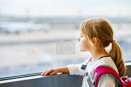 Foto de Niña en el aeropuerto esperando el embarque en la ventana grande. Cute Kid se para en la ventana contra el telón de fondo de los aviones. Mirando hacia adelante a la partida para unas vacaciones de verano familiares. - Imagen libre de derechos