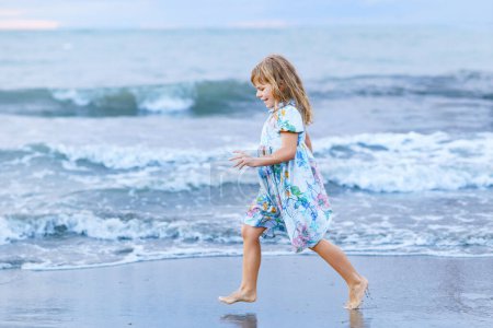 Foto de Niño feliz, niña preescolar en vestido corriendo y saltando en las olas durante las vacaciones de verano en la playa tropical exótica al atardecer. Viaje familiar en la costa del océano - Imagen libre de derechos