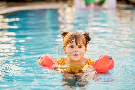 Photo for Little preschool girl with protective swimmies playing in outdoor swimming pool by sunset. Child learning to swim in outdoor pool, splashing with water, laughing and having fun. Family vacations - Royalty Free Image