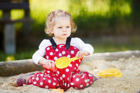 Cute toddler girl playing in sand on outdoor playground. Beautiful baby in red gum trousers having fun on sunny warm summer day. Child with colorful sand toys. Healthy active baby outdoors plays games