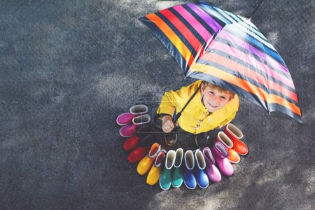 Photo for Little kid boy and group of colorful rain boots. Blond child standing under umbrella. Close-up of schoolkid and different rubber boots from high angle. Footwear for rainy fall. - Royalty Free Image