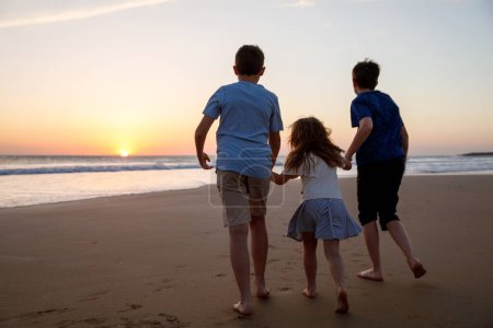 Foto de Tres niños siluetas corriendo y saltando en la playa al atardecer. familia feliz, dos chicos de la escuela y una niña preescolar. Los hermanos se divierten juntos. Vinculación y vacaciones familiares - Imagen libre de derechos
