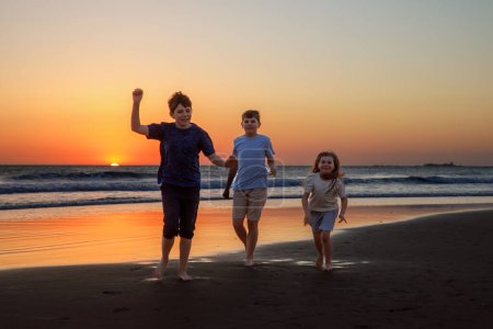 Foto de Tres niños siluetas corriendo y saltando en la playa al atardecer. familia feliz, dos chicos de la escuela y una niña preescolar. Los hermanos se divierten juntos. Vinculación y vacaciones familiares - Imagen libre de derechos