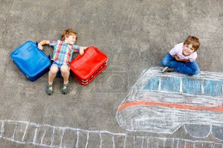 Zwei kleine Jungen amüsieren sich über die Zugbildzeichnung mit bunten Kreiden auf Asphalt. Kinder vergnügen sich mit Kreide und Buntstift-Malerei. Urlaub machen und verreisen. Freunde mit Koffern.