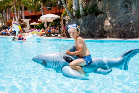 Téléchargez les photos : Joyeux petit garçon sautant dans la piscine et s'amusant en vacances en famille dans une station hôtelière. Enfant sain jouant dans l'eau avec le jouet gonflable de requin - en image libre de droit