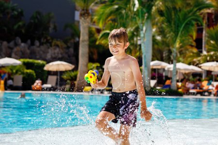 Téléchargez les photos : Joyeux petit garçon sautant dans la piscine et s'amusant en vacances en famille dans une station hôtelière. Enfant en bonne santé jouant dans l'eau avec un pistolet à eau. Rire en courant éclaboussures d'enfants
. - en image libre de droit