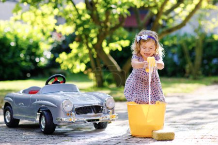 Photo for Cute gorgeous toddler girl washing big old toy car in summer garden, outdoors. Happy healthy little child cleaning car with soap and water, having fun with splashing and playing with sponge - Royalty Free Image