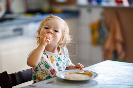 Photo for Happy little toddler girl eating delicious pancakes sitting in the kitchen. Cute child tasting different food. - Royalty Free Image