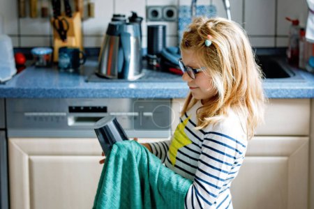 Téléchargez les photos : Petite fille essuyant la plaque avec une serviette dans la cuisine, séchant la vaisselle. enfant aidant à la maison. - en image libre de droit