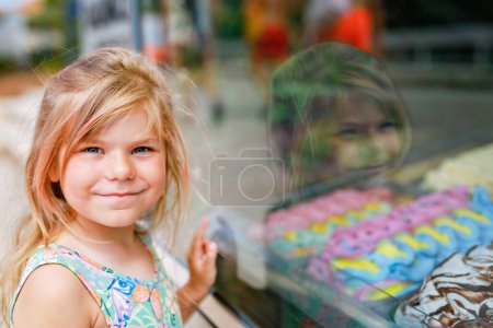 Foto de Feliz niña preescolar elegir y comprar helado en el café stand al aire libre. Lindo niño mirando diferentes tipos de helado. Dulce postre de verano en vacaciones familiares. Verano. - Imagen libre de derechos