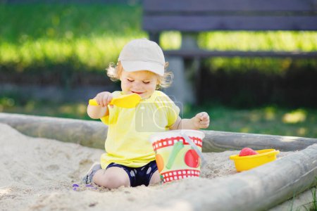 Foto de Linda niña jugando en la arena en el patio al aire libre. Hermoso bebé que se divierte en el soleado cálido día soleado de verano. Feliz niño sano con juguetes de arena y en ropa de moda colorida - Imagen libre de derechos
