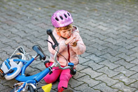 Photo for Cute little girl sitting on the ground after falling off her bike. Upset crying preschool child with safe helmet getting hurt while riding a bicycle. Active family leisure with kids - Royalty Free Image