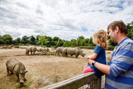 Foto de Linda adorable niña y padre viendo rinocerontes salvajes en el zoológico. Feliz bebé niño, hija y papá, familia divirtiéndose junto con animales safari parque en el cálido día de verano. Irlanda. - Imagen libre de derechos
