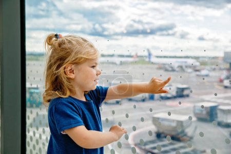 Téléchargez les photos : Jolie petite fille tout-petit à l'aéroport, voyageant. Heureux enfant en bonne santé attendant près de la fenêtre et regardant les avions. Famille en vacances d'été en avion - en image libre de droit