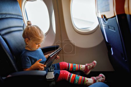 Téléchargez les photos : Petite fille en bas âge voyageant en avion. Petit enfant heureux assis par la fenêtre de l'avion et utilisant une tablette numérique pendant le vol. Voyager à l'étranger avec des enfants - en image libre de droit