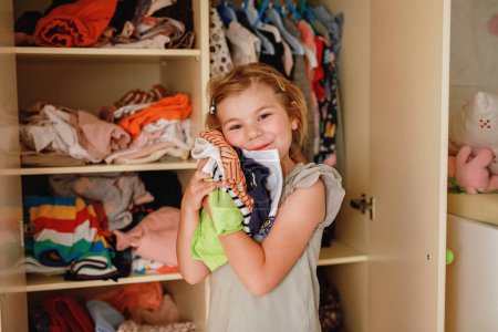 Photo for Little school girl standing by wardrobe with clothes. Preschool child putting shirts away and making decision for shirt to wear. Children get dressed in the morning for school - Royalty Free Image