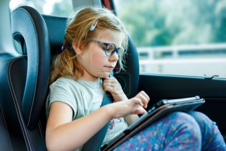 Téléchargez les photos : Petite fille joue tablette à l'intérieur de la voiture. La famille part en vacances. Enfant d'âge préscolaire s'amuser - en image libre de droit