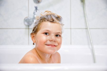 Téléchargez les photos : Enfant mignon avec mousse de shampooing et bulles sur les cheveux prenant un bain. Portrait de fille préscolaire souriante heureuse concept de soins de santé et d'hygiène. Se lave les cheveux toute seule - en image libre de droit