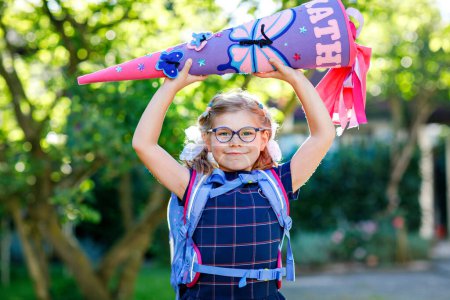 Photo for Happy little kid girl with eye glasses with backpack or satchel and big school bag or gift cone traditional in Germany for the first day of school. Healthy adorable child outdoors. Back to school. - Royalty Free Image