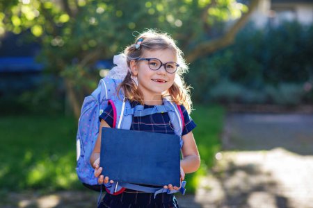 Happy little kid girl with eye glasses with backpack or satchel and big school bag on the first day of school. Healthy adorable child outdoors. Child holding chalks desk for first schoolday