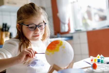 Téléchargez les photos : Petite fille peignant globe ou balle avec des couleurs. Un écolier fabriquant un globe terrestre pour un projet scolaire. Enfant heureux avec des lunettes tenant brosse - en image libre de droit