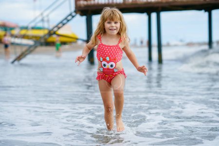 Téléchargez les photos : Enfant heureux, petite fille préscolaire en maillot de bain courant et sautant dans les vagues pendant les vacances d'été sur la plage tropicale exotique. Voyage en famille sur la côte de l'océan - en image libre de droit