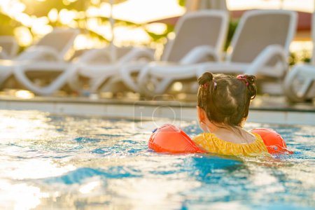 Téléchargez les photos : Petite fille d'âge préscolaire avec des natations protectrices jouant dans la piscine extérieure au coucher du soleil. Enfant apprenant à nager dans la piscine extérieure, éclaboussant d'eau, riant et s'amusant. Vacances en famille - en image libre de droit