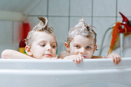 Téléchargez les photos : Frères et sœurs heureux : Deux petits jumeaux en bonne santé enfants jouent ensemble avec l'eau en prenant un bain dans la baignoire à la maison. Des gamins qui s'amusent ensemble. enfants laver les têtes et les cheveux avec un shampooing - en image libre de droit