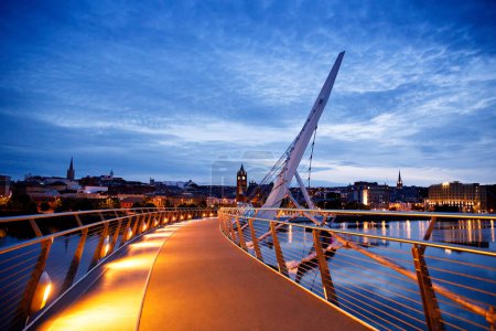 Téléchargez les photos : Derry, Irlande. Pont de la Paix illuminé à Derry Londonderry, Ville de la Culture, en Irlande du Nord avec le centre-ville en arrière-plan. Ciel nuageux nocturne avec reflet dans la rivière au crépuscule. - en image libre de droit