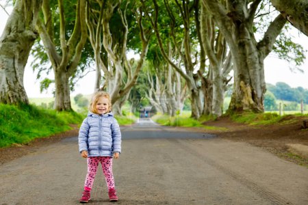 Nettes Kleinkind Mädchen zu Fuß an einem regnerischen Tag zu Beginn von The Dark Hedges. Nordirland. Glückliches Kind zu Besuch bei Eltern und Familie berühmte irische Baumallee.
