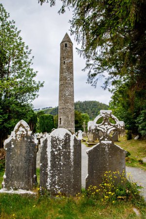Photo for Stone round tower and some ruins of a monastic settlement originally built in the 6th century in Glendalough valley, County Wicklow, Ireland on sunny day. - Royalty Free Image