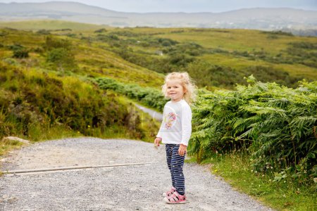 Cute little happy toddler girl running on nature path in Connemara national park in Ireland. Smiling and laughing baby child having fun spending family vacations in nature. Traveling with small kids.