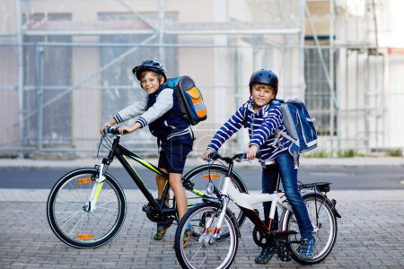 Zwei Schulkinder mit Schutzhelm fahren mit dem Fahrrad in der Stadt mit Rucksäcken. Fröhliche Kinder in bunten Klamotten radeln auf Fahrrädern auf dem Schulweg. Sicherer Schulweg für Kinder im Freien.