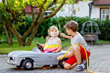 Téléchargez les photos : Deux enfants heureux jouant avec une grande vieille voiture jouet dans le jardin d'été, à l'extérieur. Enfant garçon ravitailler voiture avec petite fille tout-petit - en image libre de droit