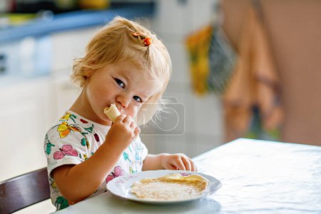Foto de Niña feliz comiendo deliciosos panqueques sentados en la cocina. Lindo niño degustación de alimentos diferentes. - Imagen libre de derechos