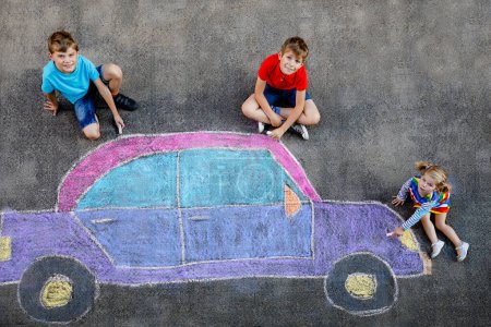 Photo for Three little children, two school kids boys and toddler girl having fun with with car picture drawing with colorful chalks on asphalt. Siblings painting on ground playing together. Creative leisure. - Royalty Free Image