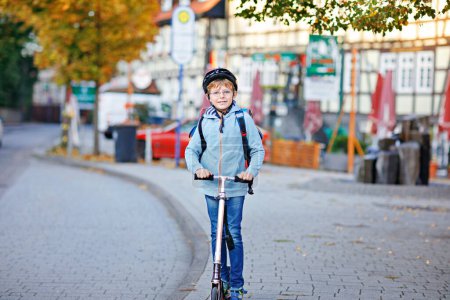 Photo for Active school kid boy in safety helmet riding with his scooter in the city with backpack on sunny day. Happy child in colorful clothes biking on way to school. Safe way for kids outdoors to school. - Royalty Free Image