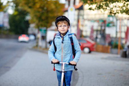 Aktive Schulkinder mit Schutzhelm fahren an sonnigen Tagen mit ihrem Roller in der Stadt mit Rucksack. Glückliches Kind in bunten Klamotten auf dem Schulweg. Sicherer Schulweg für Kinder im Freien.